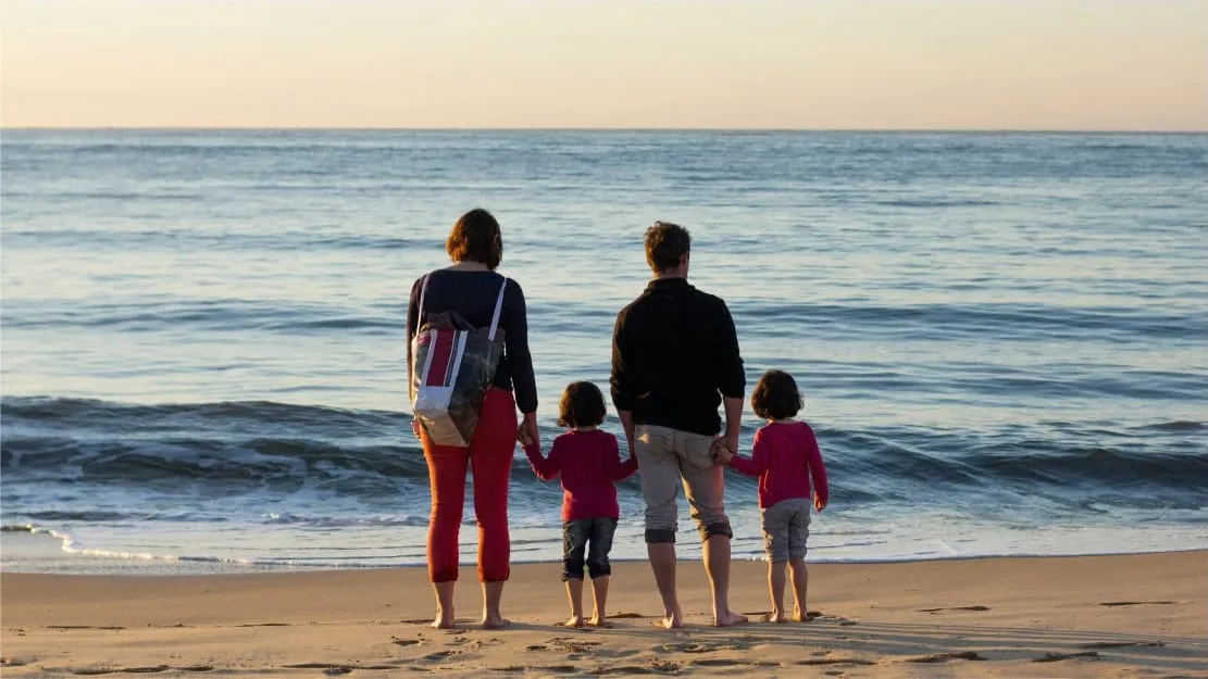 A family of four poses happily on the beach, surrounded by sand and the sparkling ocean under a clear blue sky